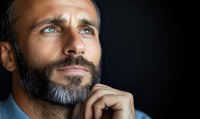Thoughtful mature man with gray beard and blue shirt looking up with hopeful expression against dark background, contemplating future possibilities.
