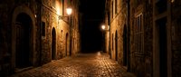 Narrow cobblestone street at night with warm glowing lanterns illuminating ancient stone buildings and arched doorways in historic European old town district.