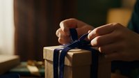Hands tying blue ribbon on kraft paper wrapped gift box with warm lighting, suitable for holiday, celebration, and gift-giving marketing materials.
