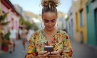 Young woman with top bun hairstyle and gold hoop earrings using smartphone on colorful street, wearing floral pattern blouse in vibrant urban setting.