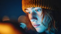 Young woman in orange knit hat illuminated by warm light against dark background with bokeh effects for winter fashion and lifestyle concepts.