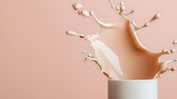 Creamy beige liquid splashing dynamically from white ceramic cup on pastel pink background, creating abstract crown-shaped splash with droplets in mid-air motion.