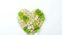 Fresh cucumber and radish slices arranged in heart shape with green lettuce leaves on white background for healthy eating and nutrition concepts.