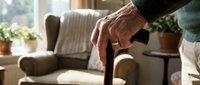 Senior person holding wooden walking cane in cozy living room with natural light streaming through window creating warm atmosphere for mobility support.