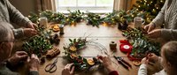 Family crafting Christmas wreaths together at wooden table with festive decorations, pine branches, ribbons and holiday supplies for seasonal DIY projects.