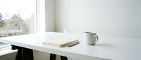 Minimalist workspace with white desk, open book, ceramic mug, and natural light from window for office and design projects.