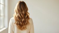 Young blonde woman with long wavy hair from behind wearing white sweater in bright modern interior with natural window light for beauty and lifestyle concepts.