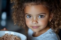 Adorable mixed-race girl with curly hair enjoying breakfast cereal, smiling at camera. Child nutrition and healthy eating habits concept for family lifestyle.
