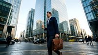 Professional businessman walking through modern financial district with leather briefcase during golden hour commute.