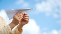 Child with light skin tone holding paper airplane against blue sky with clouds, representing childhood play, imagination, and outdoor leisure activities.