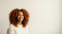 Young woman with curly reddish-brown hair, warm skin tone, wearing cream-colored long-sleeve top, smiling against neutral beige background. Portrait for lifestyle, beauty, and professional use.