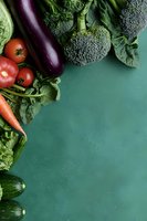 Fresh vegetables including broccoli, eggplant, tomatoes, carrots, and cucumbers arranged on green textured background with copy space for menu design.