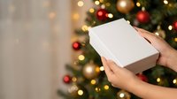 Person holding white gift box with festive decorated tree and golden lights in background, suitable for holiday shopping, seasonal promotions, and Christmas gift guides.
