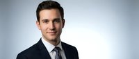 Professional young Caucasian businessman in dark suit and tie smiling confidently against neutral background for corporate headshot and business marketing.