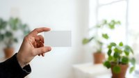 Cropped hand holding blank white card in bright room with potted plants, suitable for business cards, contact information, branding mockups, and corporate presentations.