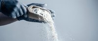Hand in dark glove pouring white powder or flour against neutral background, suitable for cooking, baking, industrial, or educational content.