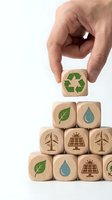 Hand placing wooden cube with recycling symbol on stack of eco-friendly blocks featuring renewable energy icons on white background.