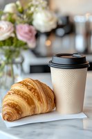 Fresh baked croissant and takeaway coffee cup on marble surface with soft focus roses in background, morning breakfast composition for cafe and bakery.