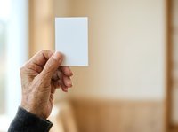 Senior adult hand holding blank white card against neutral beige wall background, suitable for business card mockups, greeting card designs, and promotional material presentations.