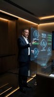 Middle-aged male professional in dark business suit presenting compliance and audit management data on holographic display in modern corporate boardroom with warm ambient lighting.