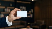 Businessman in dark suit holding blank white business card in office environment with blurred bookshelf background for corporate branding and networking purposes.