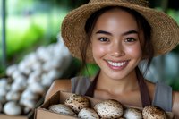 Young Asian woman in straw hat smiling while holding wooden box of fresh mushrooms, representing organic farming and local produce market concept.