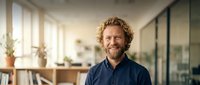 Middle-aged Caucasian businessman with curly hair and beard smiling confidently in modern office workspace with natural lighting and glass partitions.