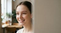 Young Caucasian woman with brown hair smiling warmly at camera in bright modern home interior with natural lighting and cozy atmosphere.