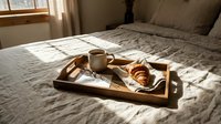 Cozy breakfast in bed with a croissant and coffee on a wooden tray, bathed in warm morning sunlight on linen bedding.