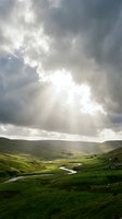 Dramatic sunlight breaks through storm clouds over rolling green hills with a winding river and rural farmhouse, capturing moody weather and pastoral landscape scenery.