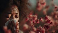 Young African American woman with curly hair blowing nose into tissue during spring allergy season with cherry blossoms in background.