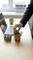 Professional male employee in business attire placing green figure into cardboard box at desk with laptop, coffee cup, and onboarding process sign for HR recruitment and hiring concepts.