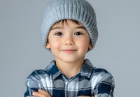 Young Asian child wearing gray knit beanie and plaid shirt smiles warmly at camera against light background, expressing joy and innocence.
