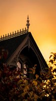 Ornate Victorian gingerbread house detail with decorative trim and finials silhouetted against warm golden sunset sky creating atmospheric architectural scene.