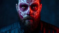Bearded man with Day of the Dead sugar skull face paint featuring roses and swirls in dramatic red and blue lighting for Halloween costume makeup design.