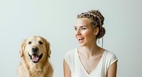 Young woman with blonde hair styled in an updo, fair complexion, wearing a white v-neck top, smiling while posing alongside a golden retriever dog against a neutral white background for pet care and lifestyle content.