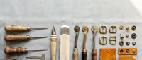Traditional leatherworking tools and supplies arranged on white background including awls, needles, thread, stamps and hardware for crafting.