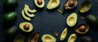 Fresh ripe avocados arranged in circular pattern on dark slate background with halved and sliced pieces showing creamy flesh and pits for healthy cooking.