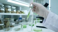 Scientist in white lab coat and gloves holding test tube with green liquid in modern laboratory setting with glass beakers and storage shelves, suitable for research, biotechnology, and medical education.