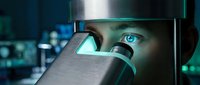 Young male scientist with blue eyes examining specimen through microscope in laboratory setting with digital displays.