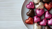 Heart-shaped chocolate candies wrapped in pink, red, and gold foil arranged on white ceramic plate against light wooden surface, romantic holiday concept.