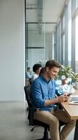 Young Caucasian male professional in blue shirt using smartphone at desk in modern office with colleagues and digital icons floating, representing mobile communication and business technology.