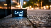 Glowing neon blue "Level up" text on dark street with bokeh lights and cobblestone pavement, shallow depth of field creating atmospheric gaming motivation scene.