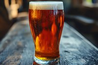 Amber craft beer in pint glass on rustic wooden bar counter with foam head and bubbles, backlit by warm lighting creating rich golden hues and texture.