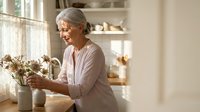 Senior Caucasian woman arranging dried flowers in ceramic vase at home kitchen counter, peaceful domestic lifestyle moment for wellness content.