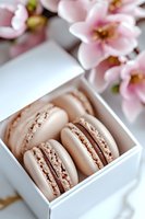 Delicate French macarons in soft beige color with chocolate filling arranged in white gift box, decorated with pink cherry blossom flowers in background.