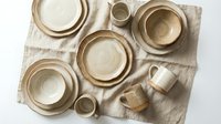 Handmade ceramic dinnerware set in neutral beige tones arranged on linen fabric, featuring plates, bowls and mugs for rustic kitchen and dining decor.