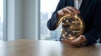 Businessman in dark suit protecting golden cryptocurrency coin with circuit board pattern between hands at office desk near window.