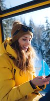 Young Caucasian woman in yellow ski jacket and knit hat using smartphone in cable car gondola during winter mountain vacation with snowy forest view.
