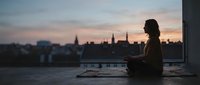 Woman sitting by window overlooking river and city skyline at dusk, contemplative mood, urban solitude, suitable for lifestyle and editorial use.